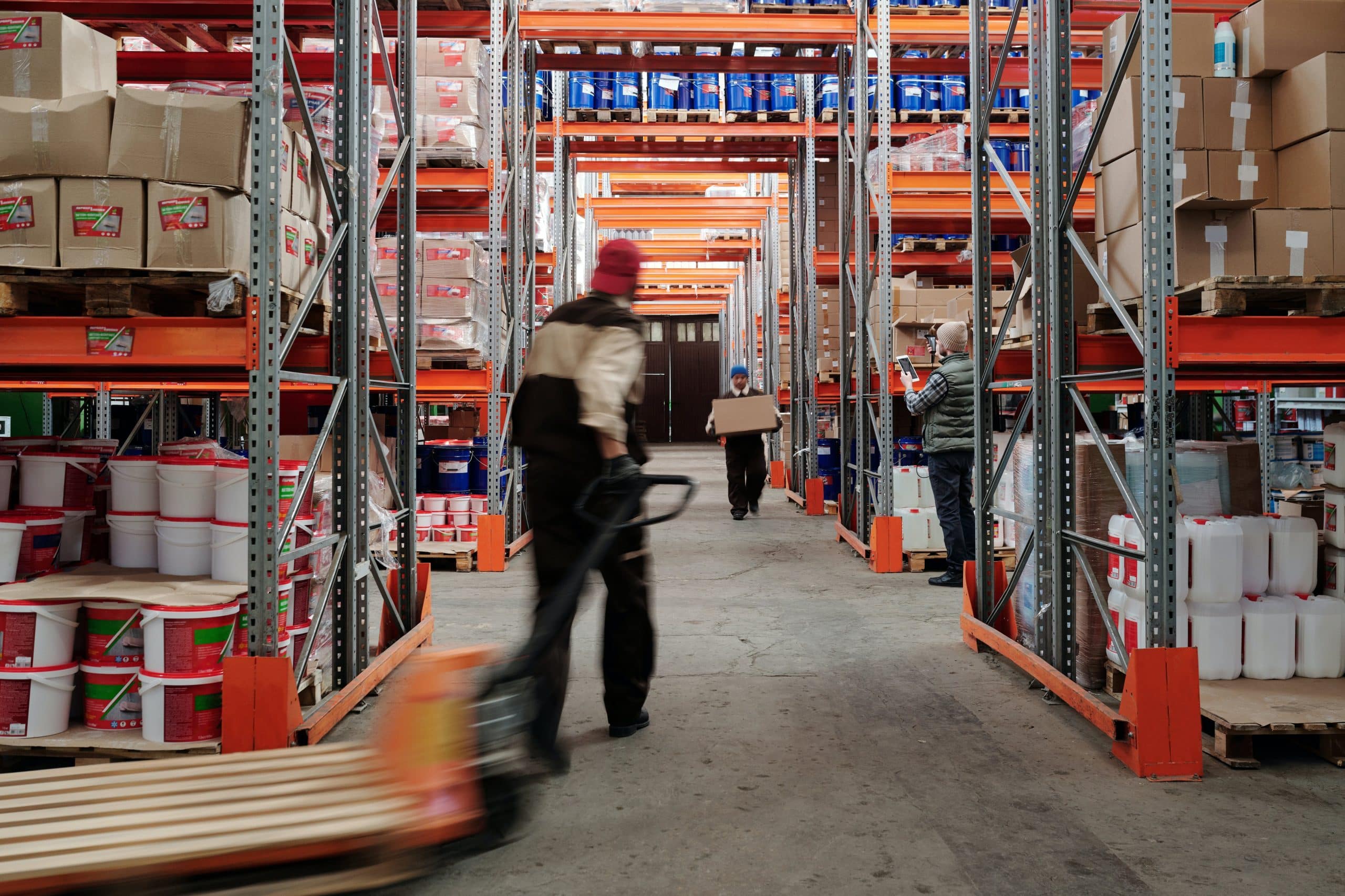 A bustling warehouse with workers engaged in various tasks, such as moving goods with a pallet jack and scanning items, amidst tall storage racks filled with inventory.